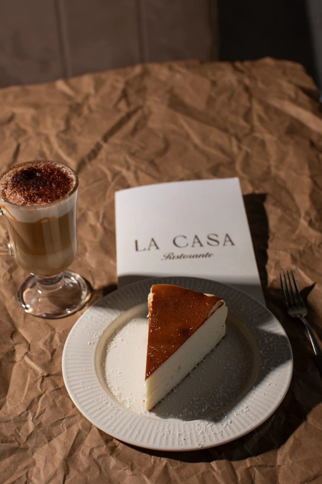 A slice of cake, a glass of coffee, and a menu on a textured brown tablecloth.