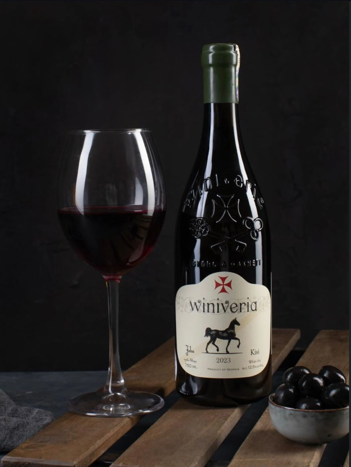 A wine bottle and a filled wine glass with dark grapes against a dark backdrop.
